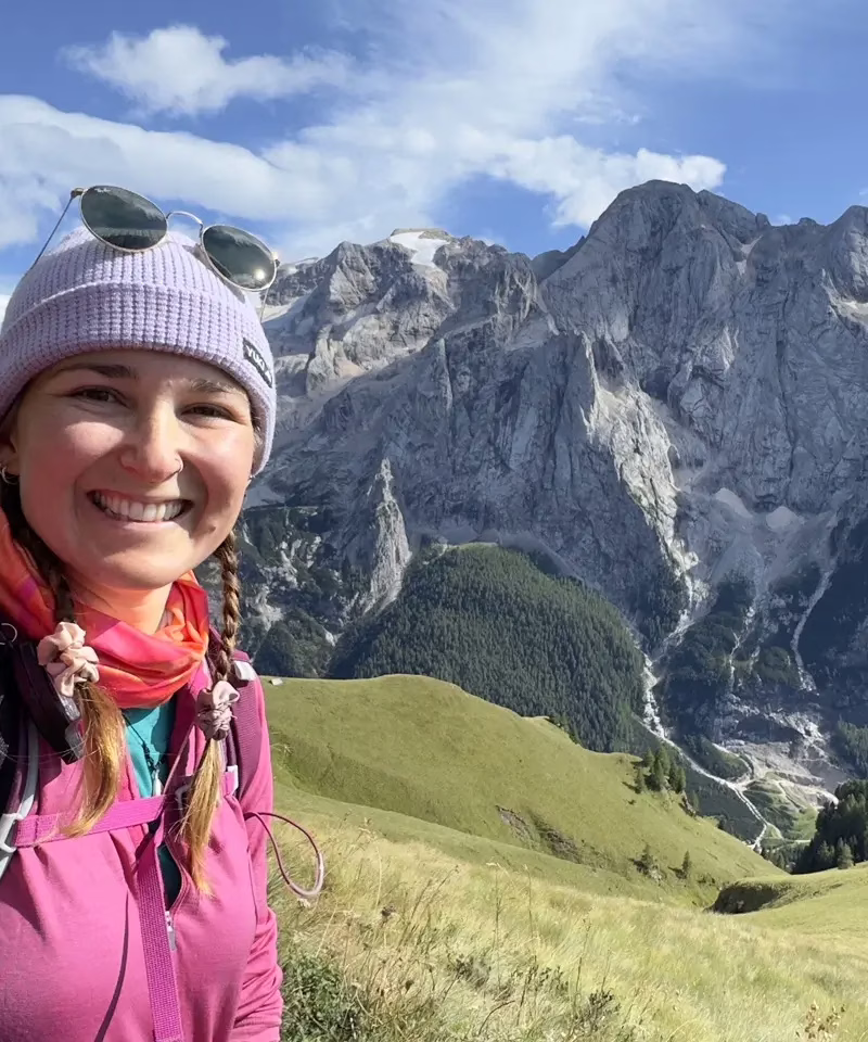 Selfie taken of myself in front of a mountain range in the Dolomites in Italy with green rolling hills and jagged granite peaks in the background.